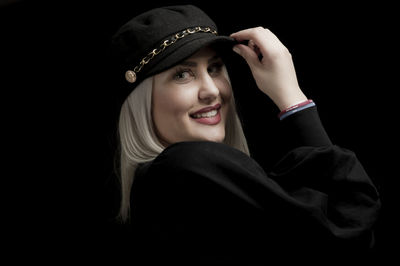 Portrait of smiling young woman wearing cap against black background