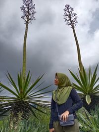 Woman wearing hijab standing amidst plants against cloudy sky