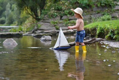 Happy little boy in yellow rain boots playing and fishing with ship boat in lake