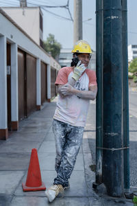 Full length of man in safety hardhat leaning on lamp post and talking on mobile phone on sidewalk