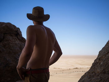 Rear view of shirtless man standing at rock against clear sky, namib naukluft national park, namibia