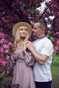 Portrait man and a woman in hat stand by a blooming pink cherry tree in summer