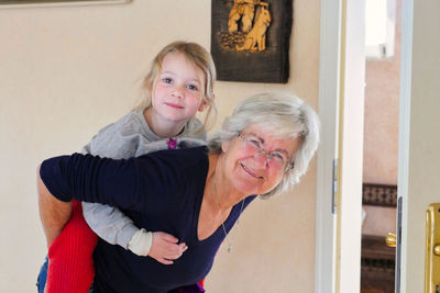 Portrait of smiling grandmother carrying grand daughter on back at home