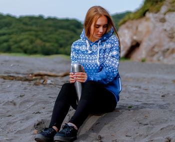 Young woman holding insulated drink container while sitting at beach