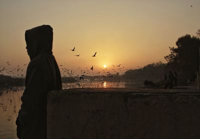 Side view of man standing with birds flying over river during sunrise