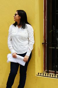 Woman holding white fabric while leaning on yellow wall by window