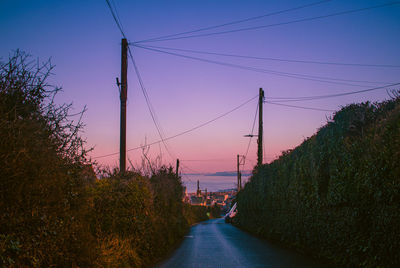 Road by electricity pylon against sky during sunset