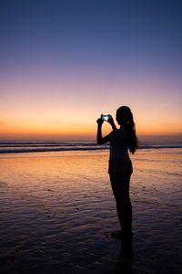 Silhouette woman photographing sea against sky during sunset