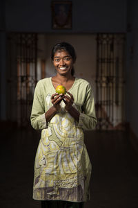 Portrait of smiling woman holding orange while standing at home