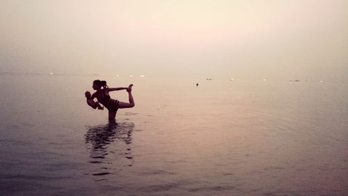 Woman holding baby and doing yoga in water at sea during sunset