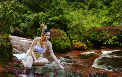 Full length of woman in traditional clothing sitting by stream in forest