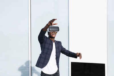 Smiling young man wearing vr glasses gesturing in front of wall