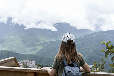 Rear view of young woman in cap with backpack looking at view of caucasian mountains and cloudy sky 