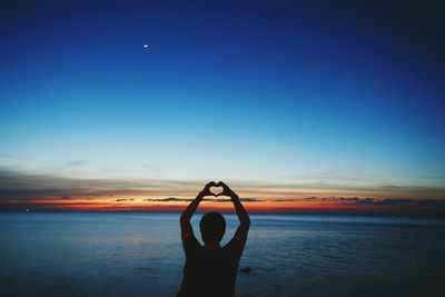Silhouette man making heart shape while standing at beach against blue sky during sunset