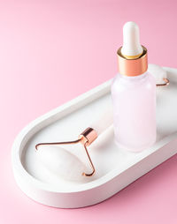 Cosmetic serum in glass bottle and face roller on white concrete tray on pink background. skincare