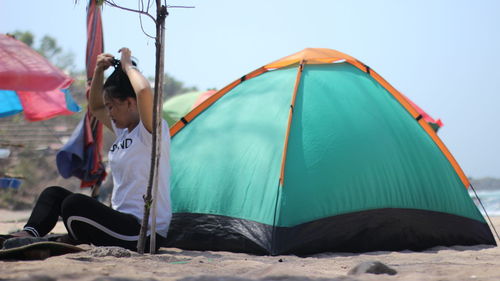 Side view of woman sitting by tent on beach against clear sky
