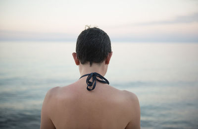 Rear view of shirtless woman standing at beach against sky during sunset