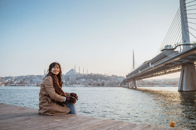 Portrait of woman sitting on pier over river against clear sky