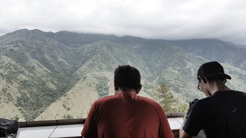 Rear view of friends with camera enjoying view of mountains against cloudy sky