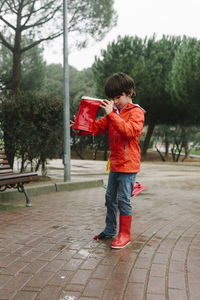 Curios kid pouring rain water from red rubber boot in wet park near wooden bench in gray day