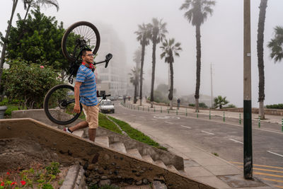 Side view of latin male carrying bicycle while walking on steps on street with green trees and pathway on foggy day