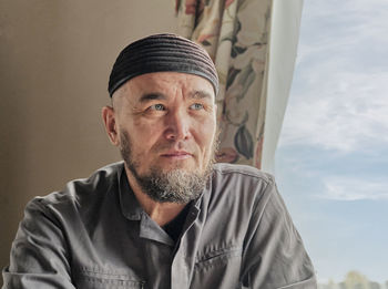 Senior asian man with beard wearing skullcap sitting by window and looking into distance. 