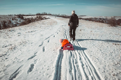 Rear view of parent pulling sled with child on snow covered field