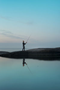 Man fishing in sea while standing against sky during sunset