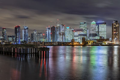 Illuminated buildings by river against sky in city