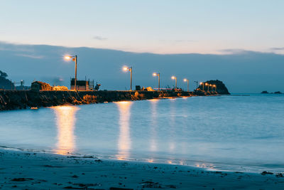 Illuminated factory by sea against sky at sunset