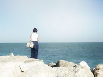 Rear view of woman standing at rocky shore against clear sky