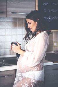 Side view of pregnant young woman holding mug while standing in kitchen