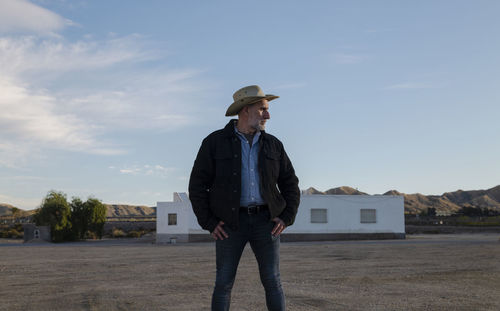 Adult man in cowboy hat standing in front of white building against sky