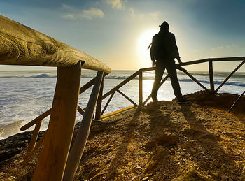Man standing on railing by sea against sky during sunset