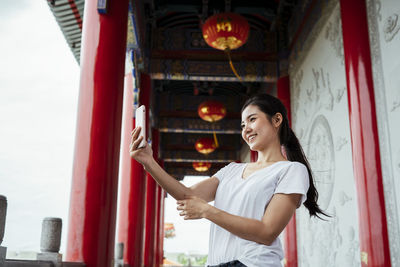 Smiling young woman using smart phone standing against wall