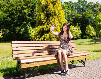 Young woman taking selfie while smart phone while sitting on bench against trees in park