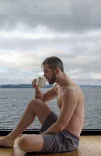 Side view of shirtless man having coffee while sitting by window against sea