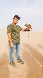 Full length of man standing on sand with eagle on hand against sky