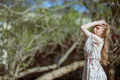 Side view of woman with hand in hair standing against trees at park