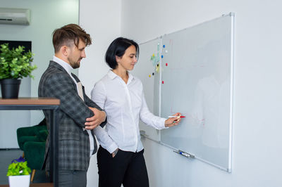 A man and a woman in formal suits are standing in the office near a blackboard with notes