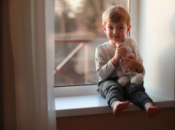 A little smiling boy sits on a windowsill in the evening light and hugs a teddy bear. 