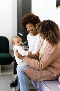 Mother taking daughter on lap while aunt sitting beside on sofa at home