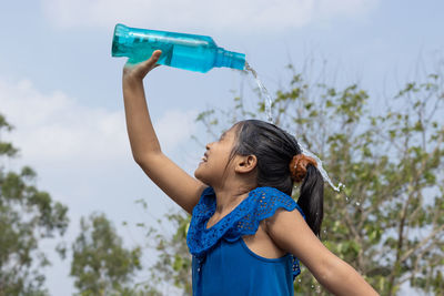 An indian girl pouring water on head from a blue plastic bottle under sky to cool down in summer