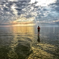 Boy standing in sea against sky during sunset