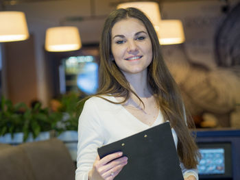 Portrait of confident young woman with clipboard standing in restaurant