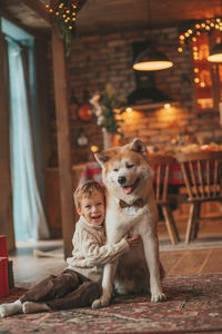 Candid authentic happy little boy in knitted beige sweater hugs dog with bow tie at home on xmas