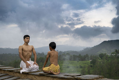 A young handsome boy doing yoga with a kid on the roof of a house situated in the middle of mountain