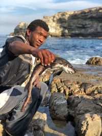 Smiling man catching fish while sitting on rock against sea at beach