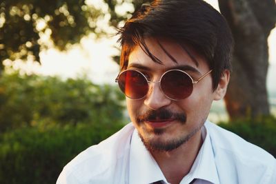 Close-up of young man wearing sunglasses against tree during sunset