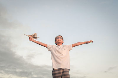 Low angle view of boy with arms outstretched holding paper airplane against sky